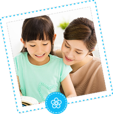 A caring adult woman gently leans over to help a smiling young girl with a book, representing a supportive and nurturing learning environment. The image is framed with a dashed-line border and a small flower icon at the bottom.